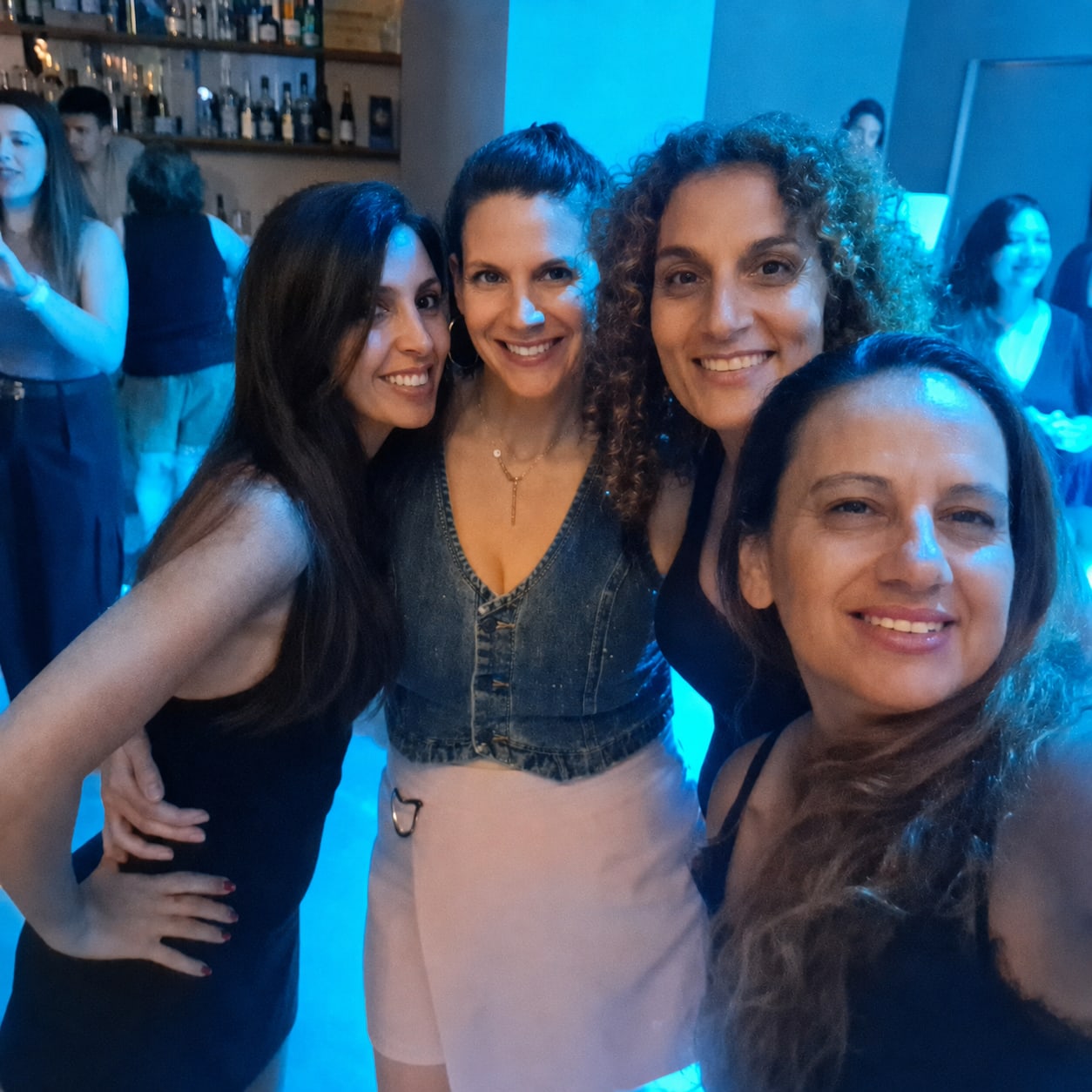 ארבע חברות מצטלמות יחד ליד הבר, הרחבה מאחוריהן בתאורה כחולה