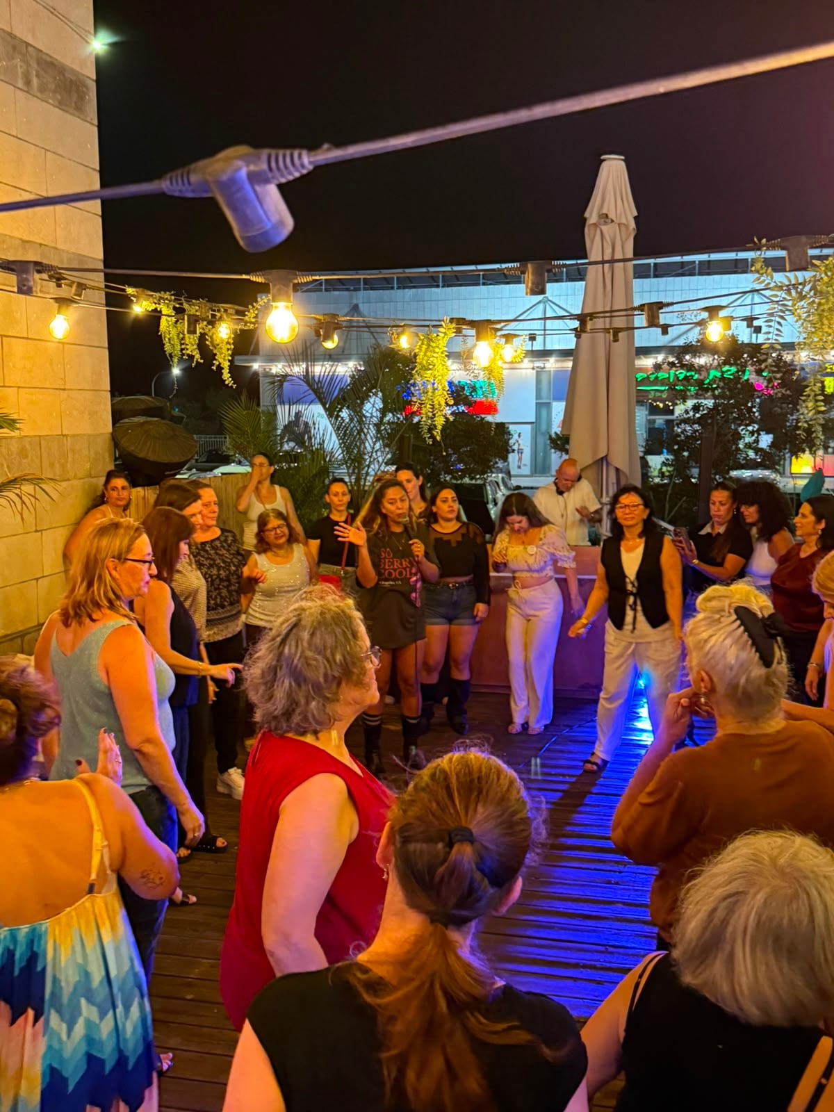 מעגל נשים על הרחבה, שתיים במרכז מובילות את הריקוד, שולחנות ואורות מאחור