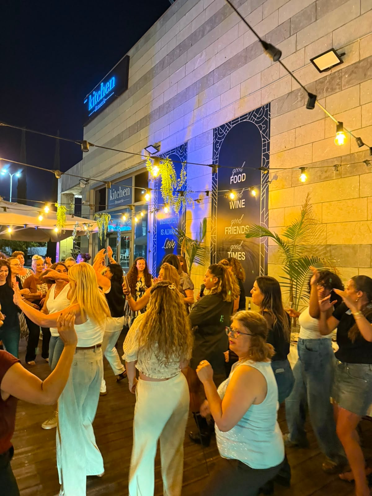 המתחם בערב, שרשראות אורות ושלט של המקום, ונשים רוקדות לפניו